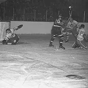 Ice-level during action of a hockey game. A goal-tender reaches down with his blocker to stop a puck. An opposition forward and two of his own players are in front of his net.